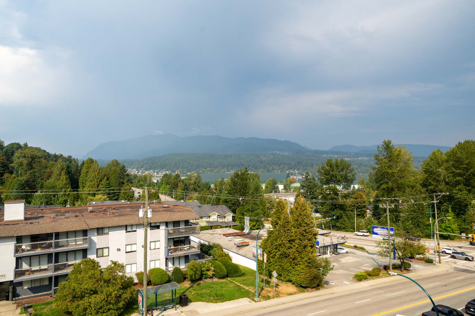 View of a residential area with low-rise buildings, trees, a road with traffic, and forested mountains in the background under a cloudy sky.