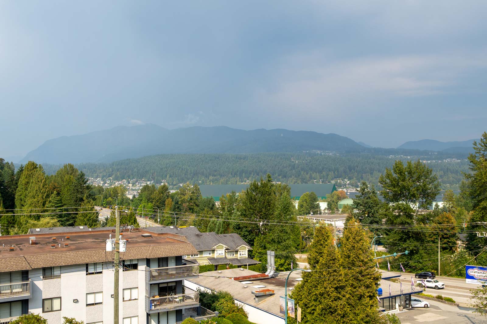 View of a residential area with apartment buildings, roads, and trees in the foreground, and a lake, forest, and mountains in the background under a partly cloudy sky.