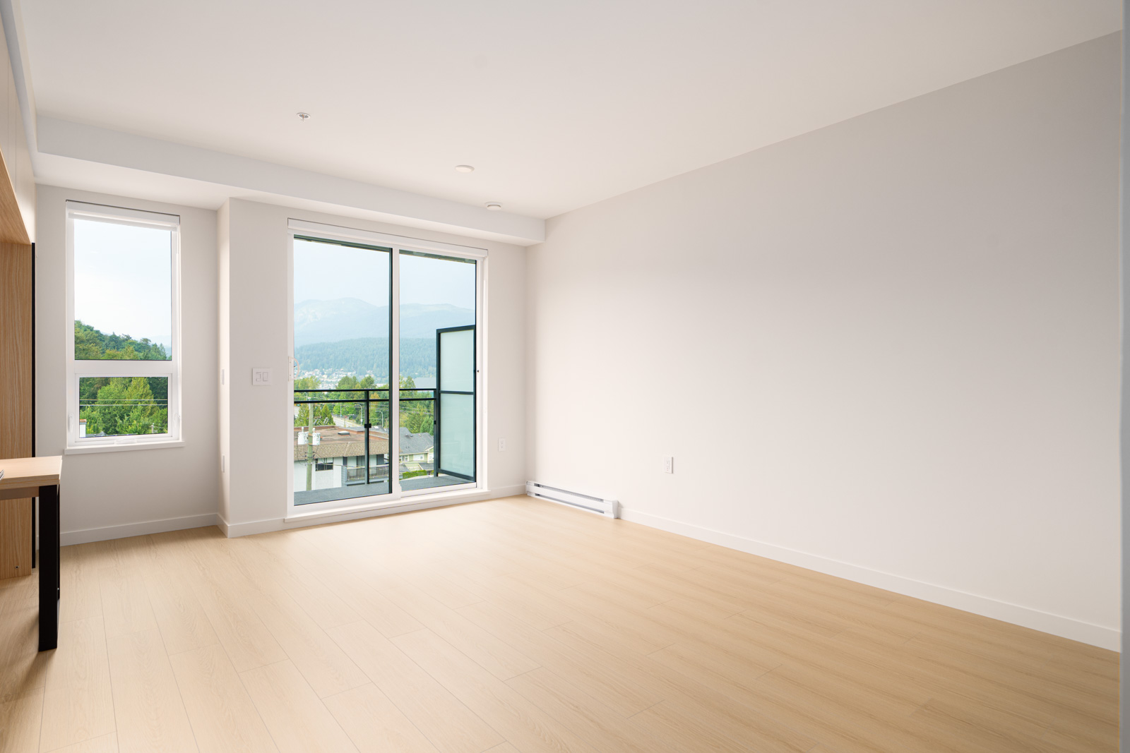 Unfurnished room with light wood flooring, white walls, large windows, and a sliding glass door leading to a balcony with a view of trees and mountains.