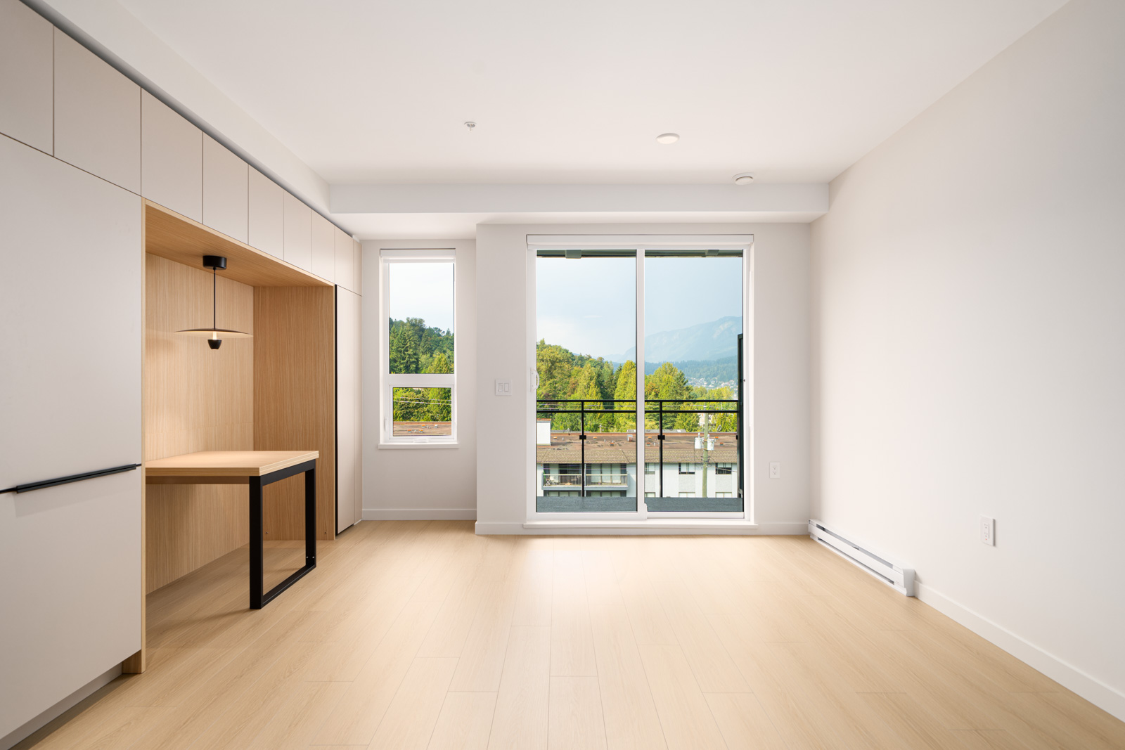 A bright, empty room with light wood flooring, built-in desk, large window, and glass doors opening to a balcony with a view of trees and mountains.