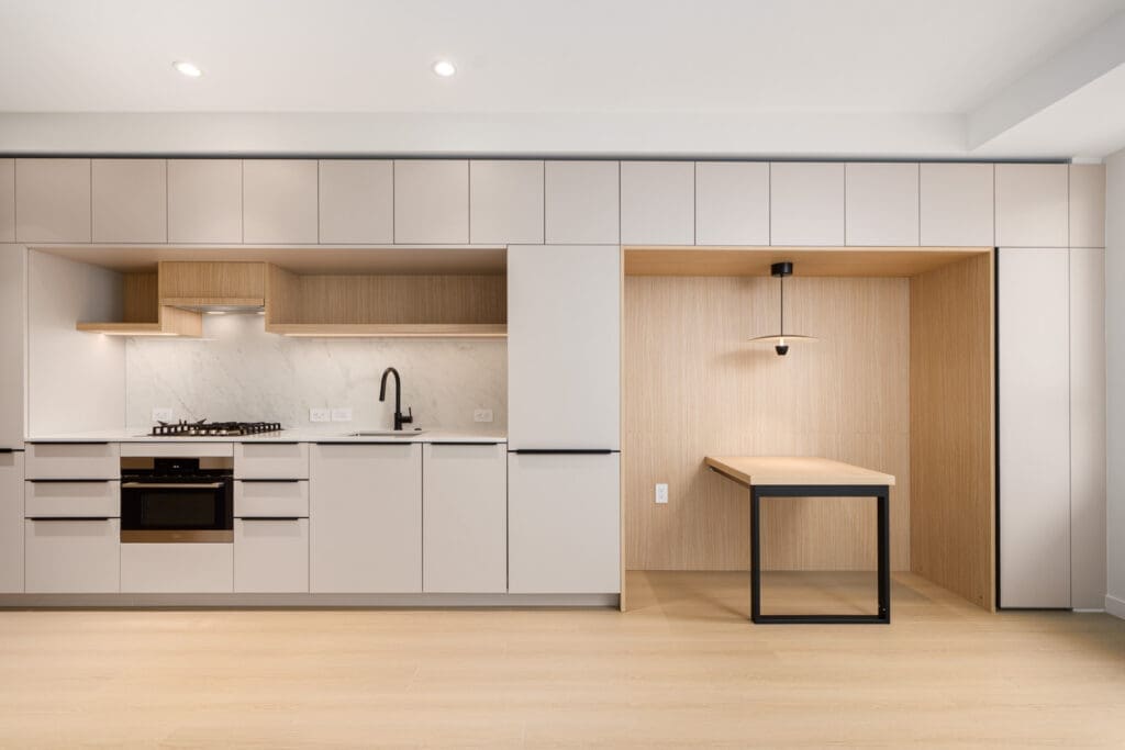 Modern kitchen with light wood and white cabinetry, integrated appliances, a black faucet, and an adjacent built-in desk area with a pendant light.