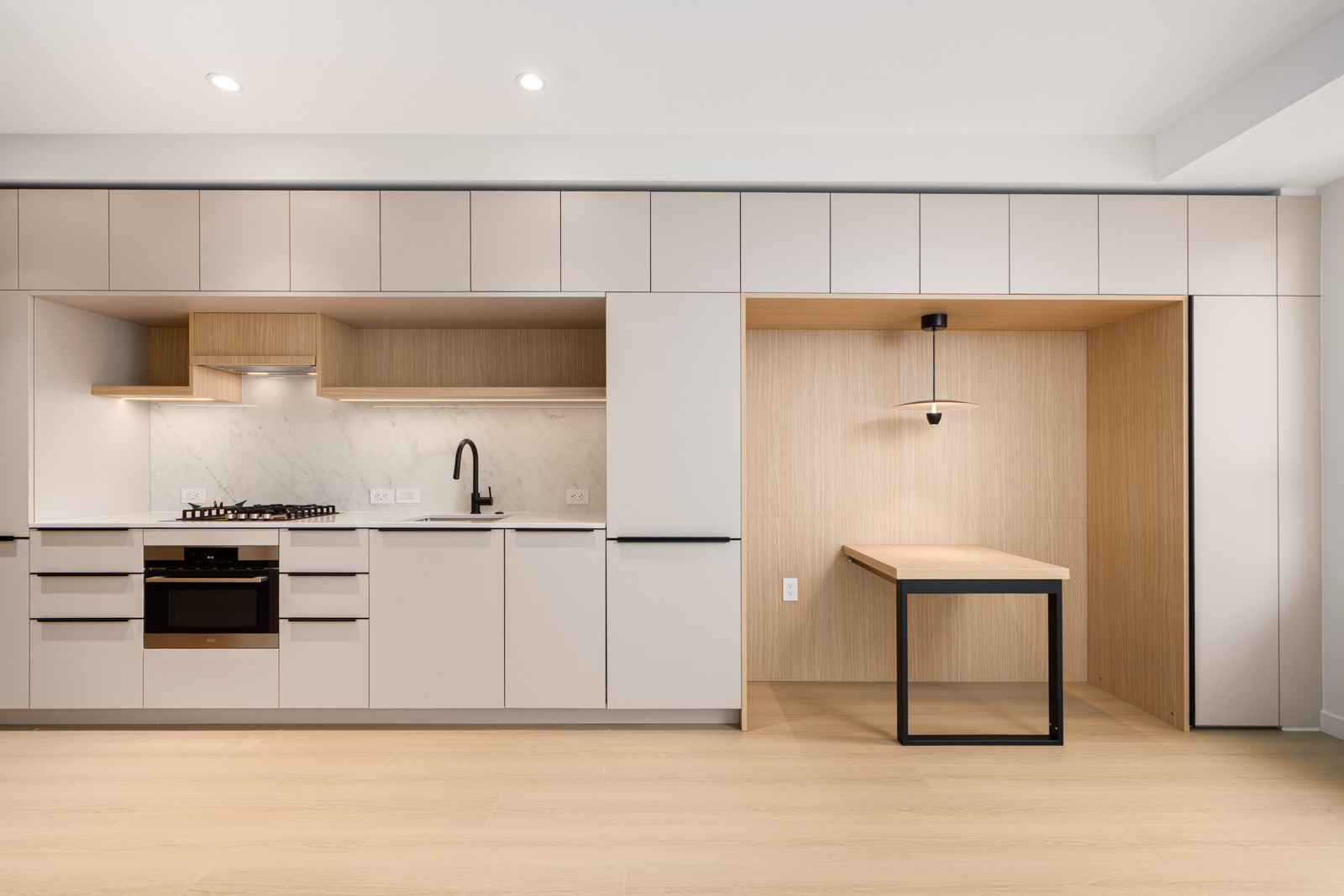Modern kitchen with light wood and white cabinetry, integrated appliances, a black faucet, and an adjacent built-in desk area with a pendant light.