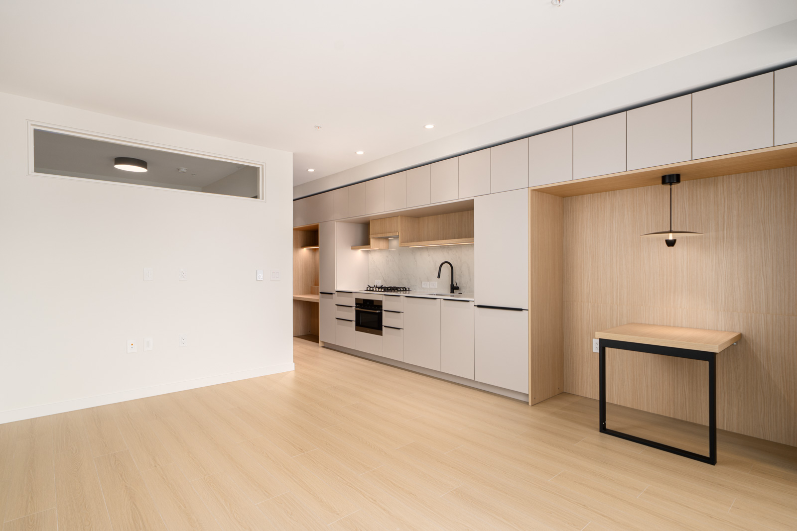 Modern minimalist kitchen with light wood flooring, built-in appliances, beige cabinets, and a small built-in desk area with a wall-mounted lamp.