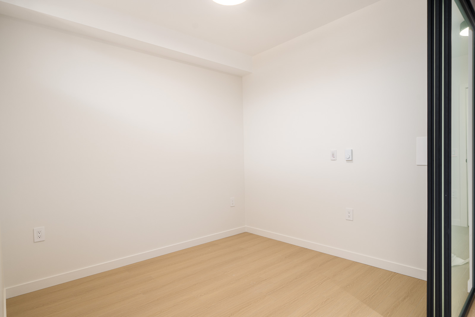 A small, empty room with white walls, a light wood floor, ceiling light, electrical outlets, and a glass door partially visible on the right side.