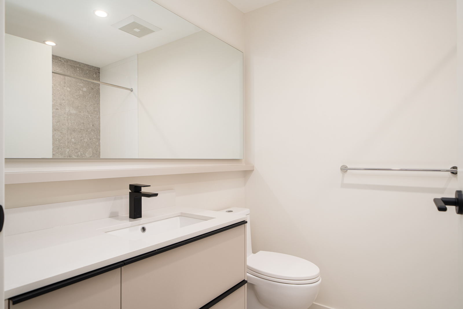 A modern bathroom with a white vanity, black faucet, large mirror, toilet, towel bar, and light-colored walls.