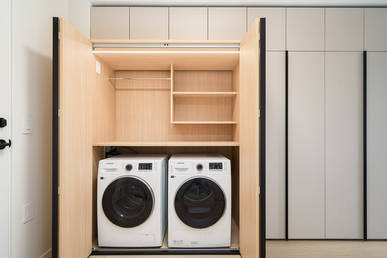 A front-loading washer and dryer are installed side by side in a built-in wooden laundry closet with shelves and hanging space above.