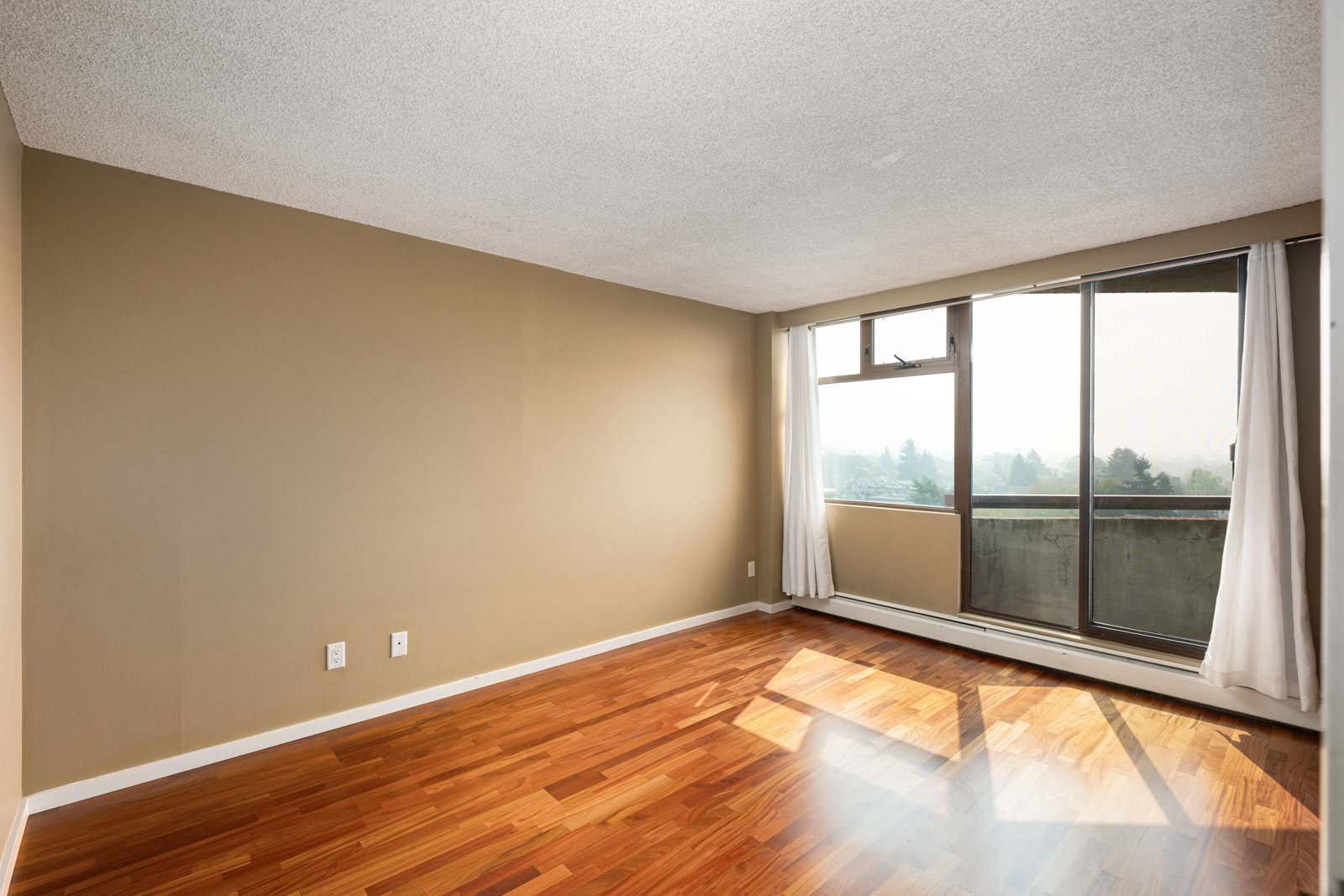 Empty room with light brown walls, wood flooring, and a large window with white curtains leading to a balcony with a view of trees.
