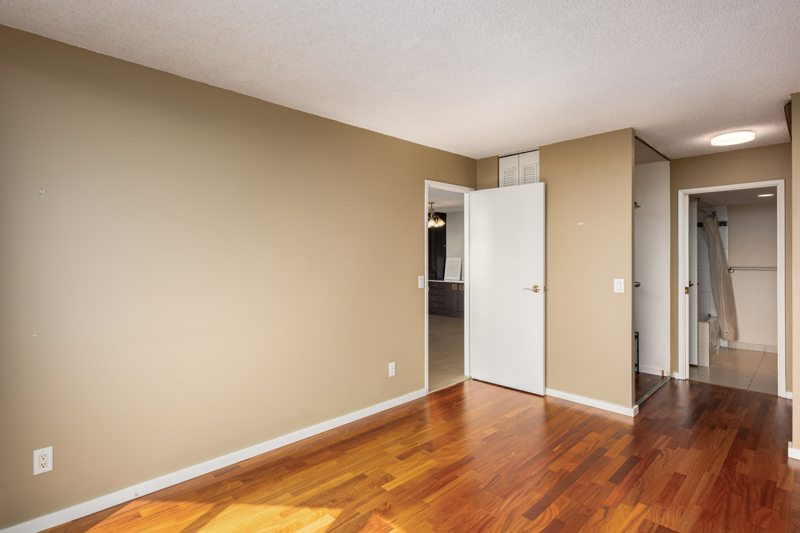 Empty room with tan walls, white doors, and wood flooring; view into an adjoining hallway and bathroom with visible shower curtain.