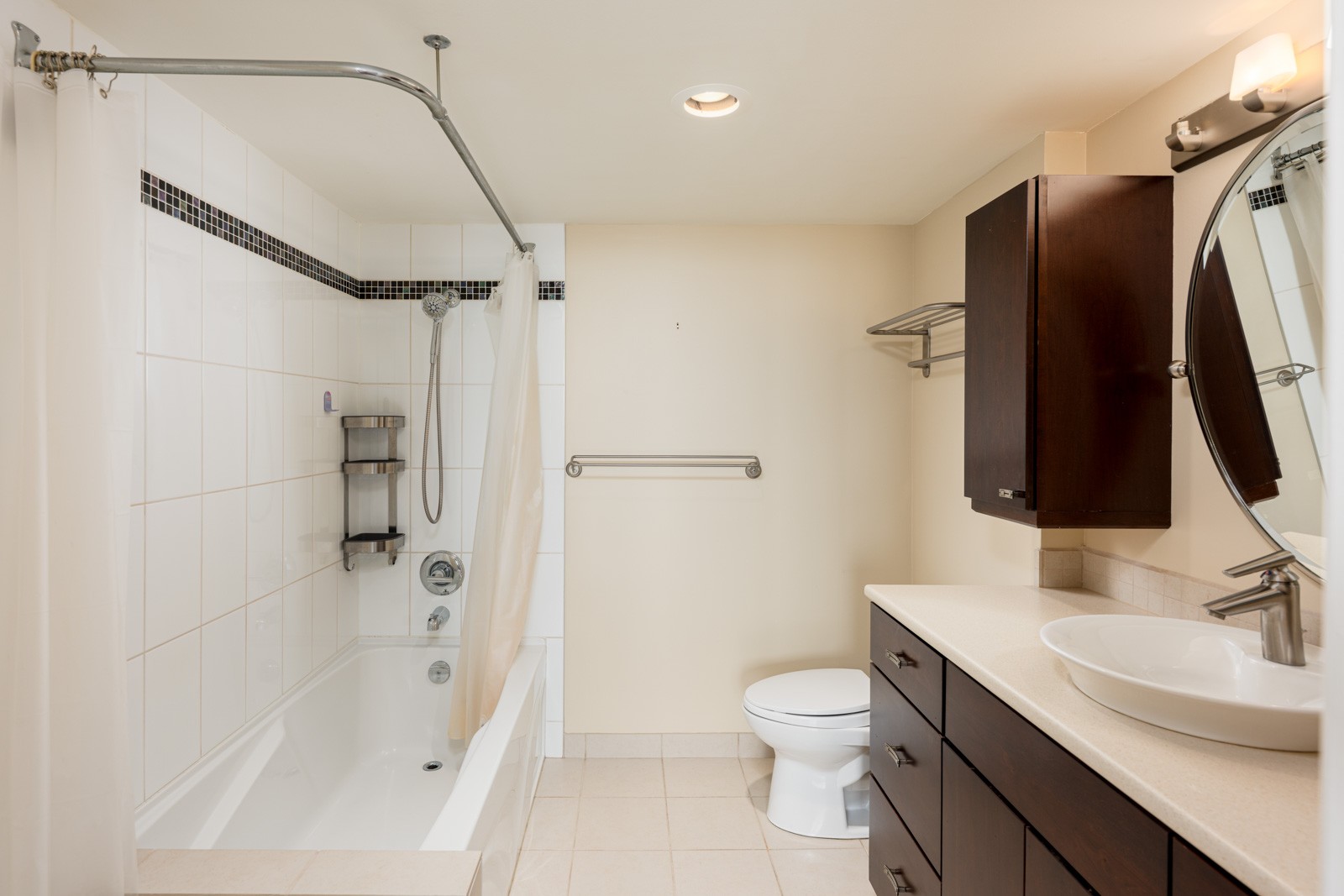 Modern bathroom with a bathtub and shower, white tiles, dark wood cabinets, a round mirror, countertop sink, toilet, and a towel bar on a beige wall.