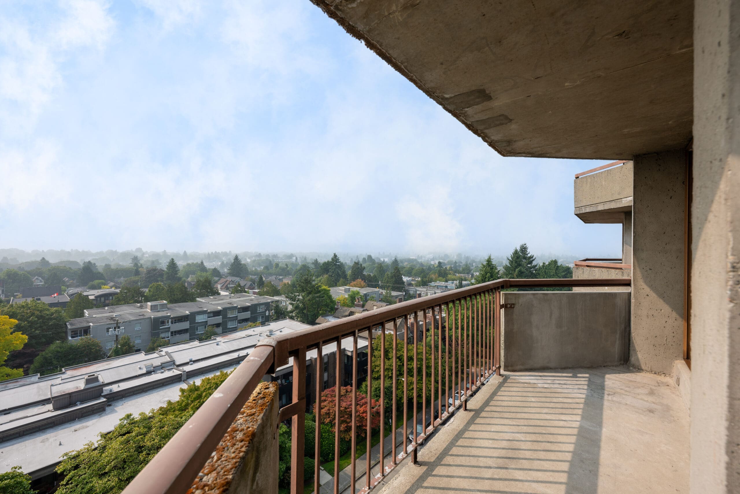 A concrete balcony with a metal railing overlooks a suburban neighborhood with trees and low-rise buildings under a cloudy sky.
