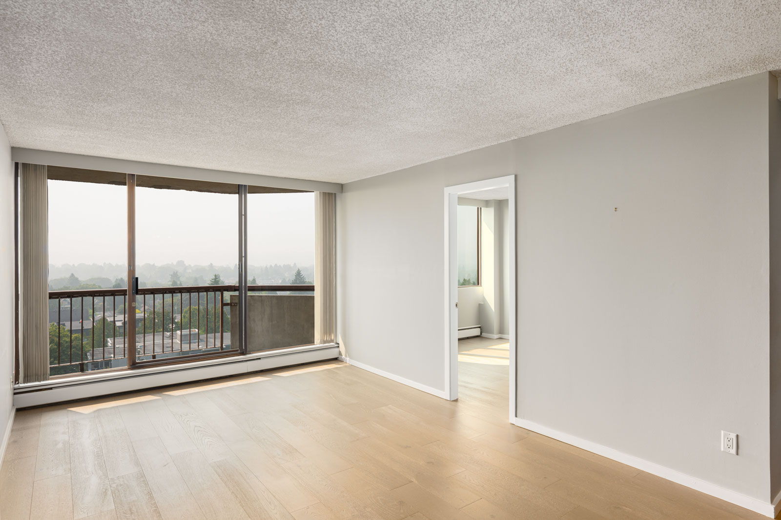Unfurnished room with light wood flooring, large window and sliding door to a balcony, and an open doorway leading to another bright room.