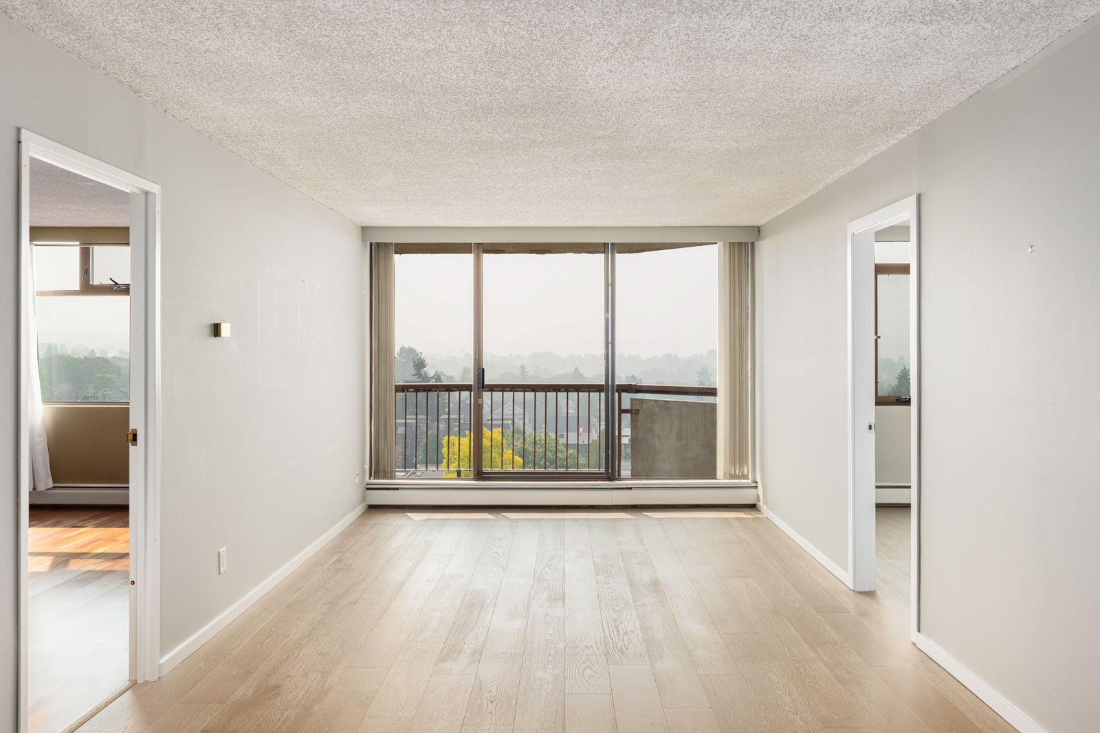 Empty, unfurnished living room with light wood flooring, white walls, and large sliding glass doors opening to a balcony with a view of trees and distant landscape.