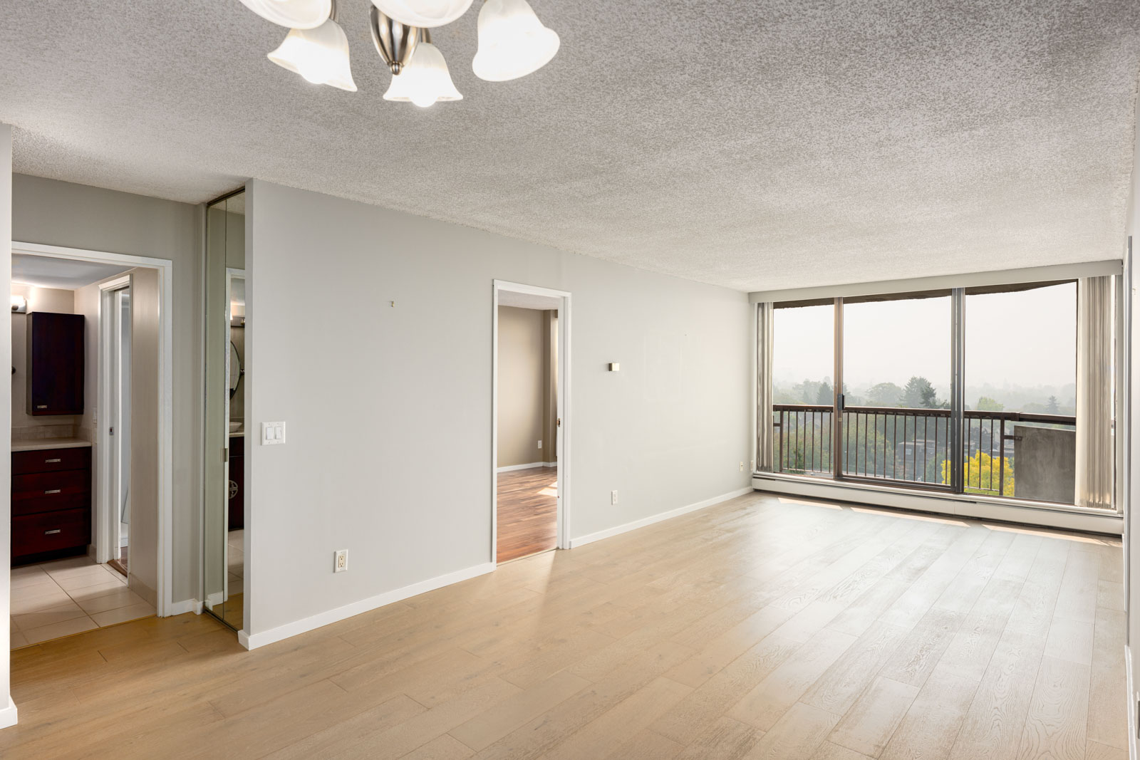 Bright, empty living room with large windows, light wood floors, and a view of trees outside. A bathroom and another room are visible to the left.