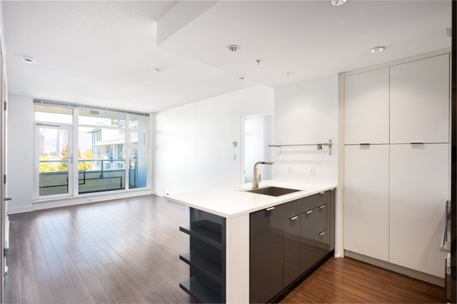 Modern, unfurnished open-plan apartment with a kitchen featuring a sink island, white cabinets, wood flooring, and large windows leading to a balcony.