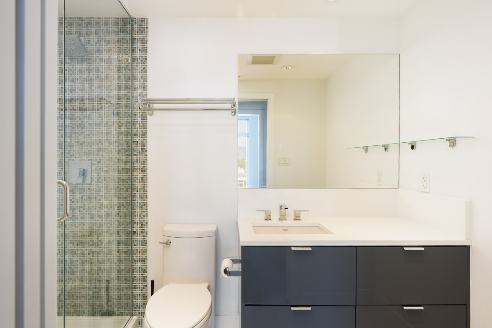 Modern bathroom with a glass-enclosed shower, mosaic tile wall, toilet, dark vanity with sink, large mirror, and glass shelf above the sink.