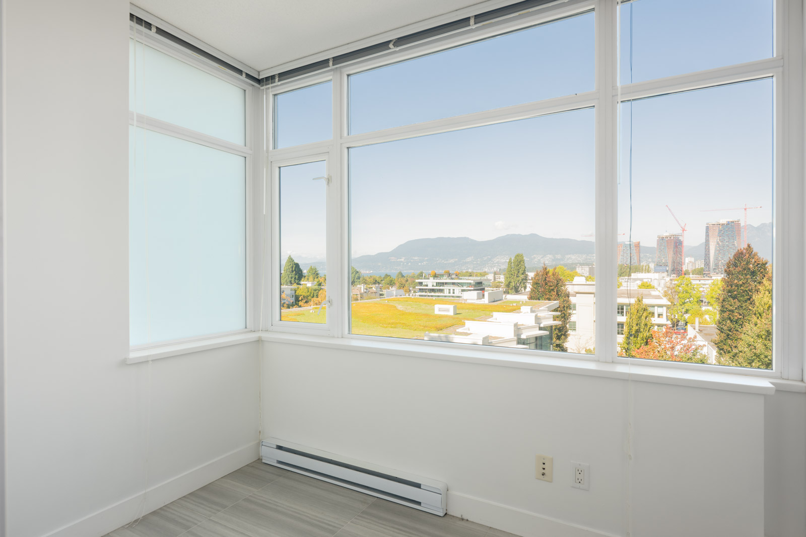 A bright, empty corner of a modern room with large windows, white walls, tile flooring, and a view of buildings, trees, and mountains in the distance.