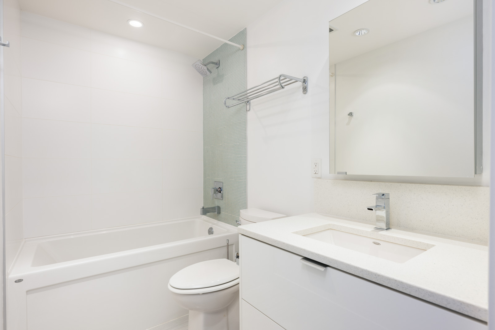 Modern bathroom with a white bathtub, toilet, sink with a rectangular mirror, and a wall-mounted towel rack. The space is brightly lit with minimalist design.