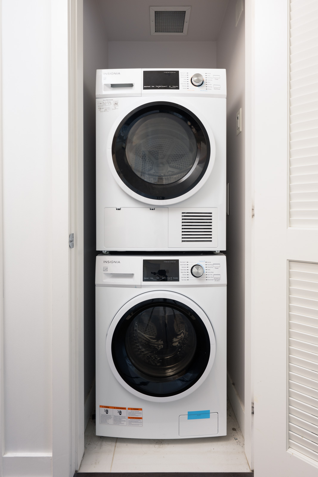 A stacked white washer and dryer set is installed in a narrow laundry closet with louvered doors.