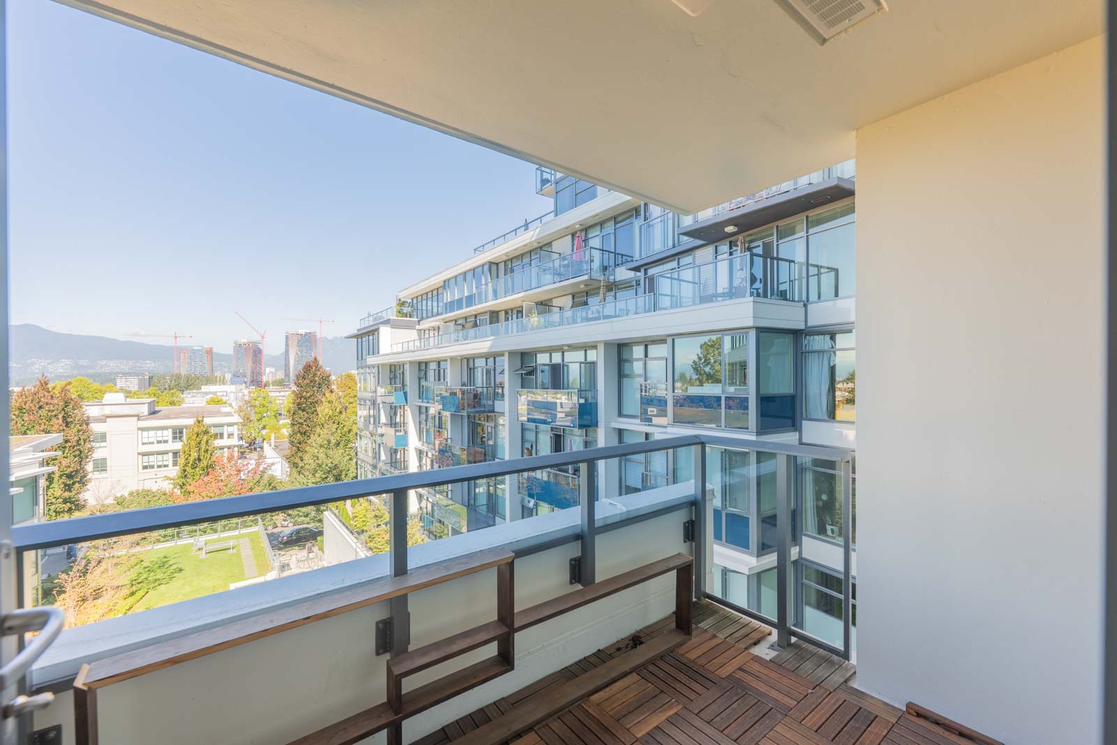 View from a modern apartment balcony with glass railings, overlooking adjacent buildings, greenery, and distant construction cranes under a clear sky.