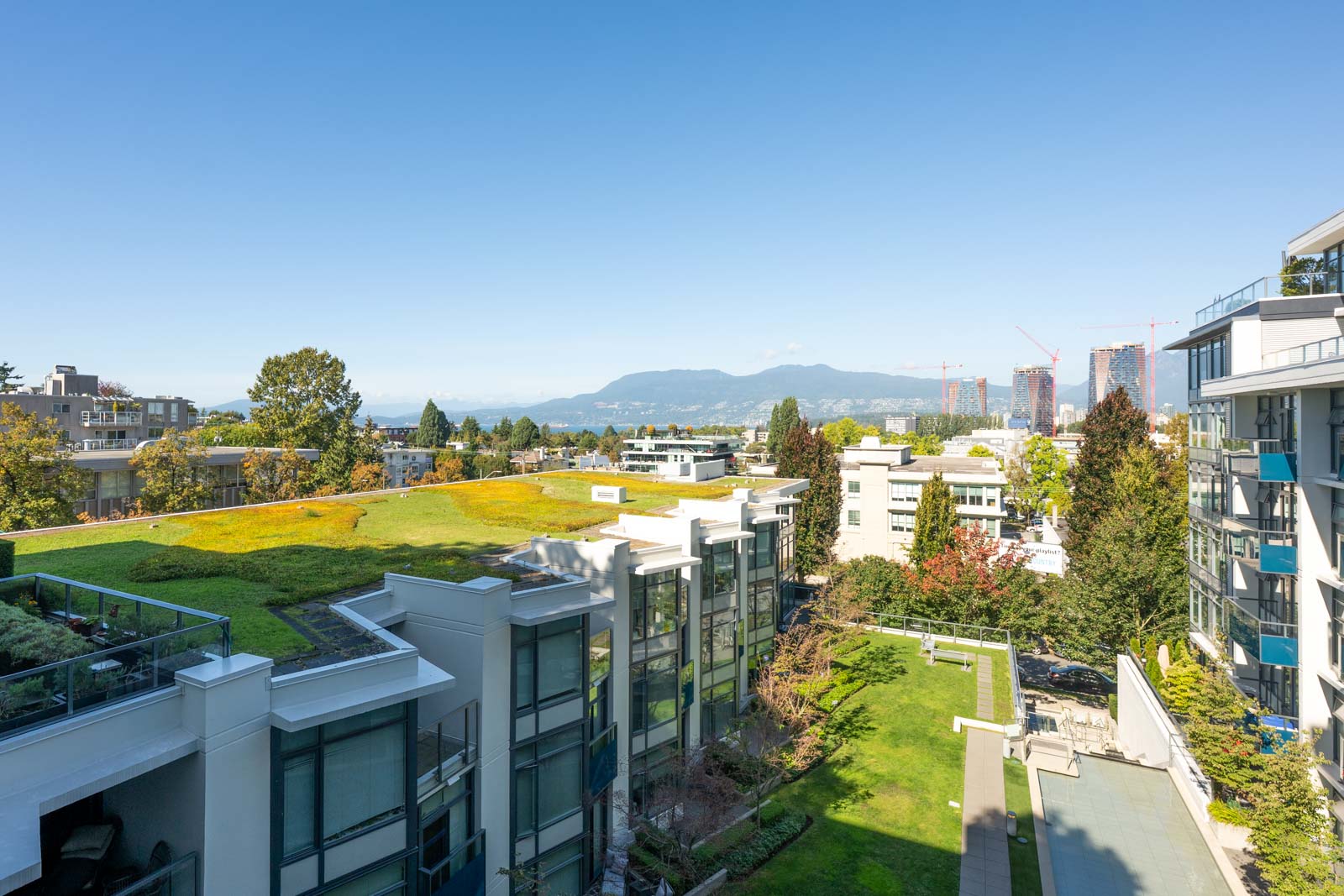 View of modern apartment buildings with green roofs, surrounded by trees, under a clear blue sky with mountains visible in the background.