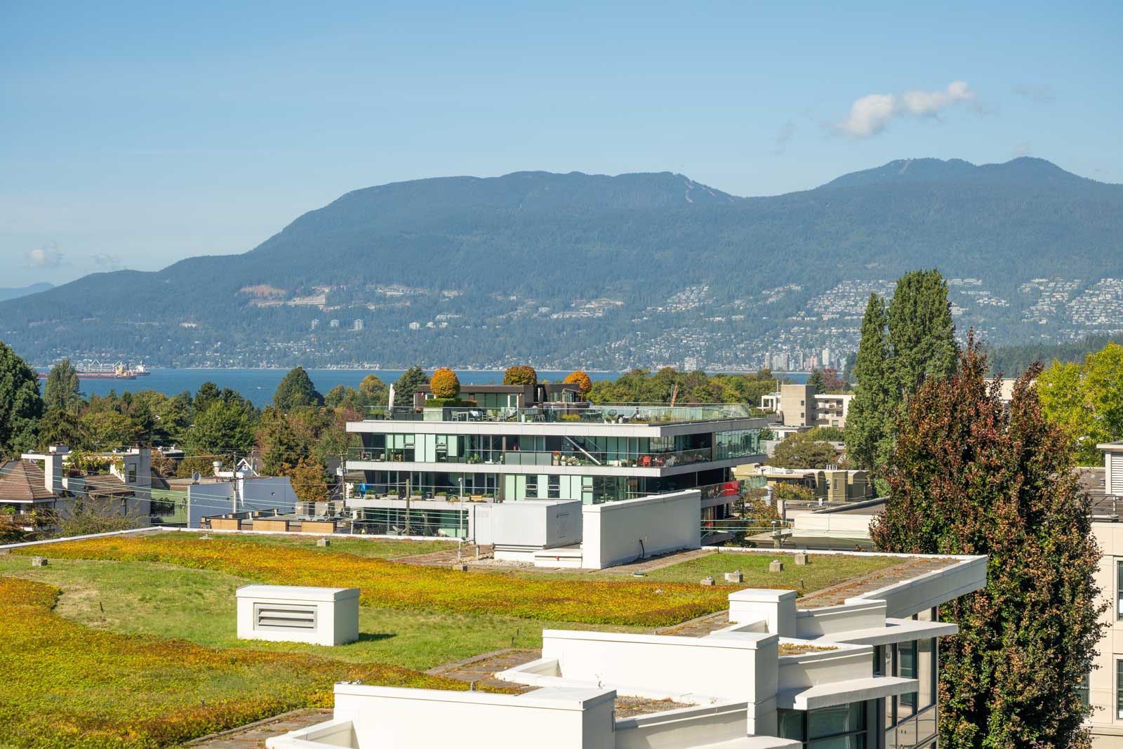 Modern residential buildings with green rooftops are set against a backdrop of mountains, trees, and a blue sky on a sunny day.