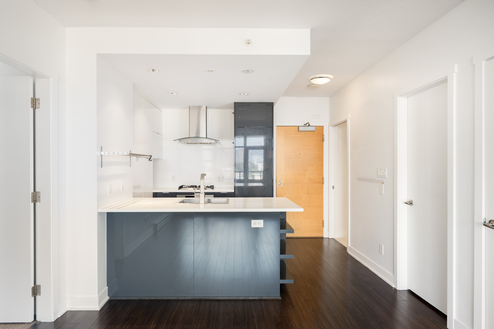 Modern kitchen with white walls, dark wood flooring, a light wood front door, and a gray-blue island with a sink and stovetop. Three doors are visible; one is open showing a closet.