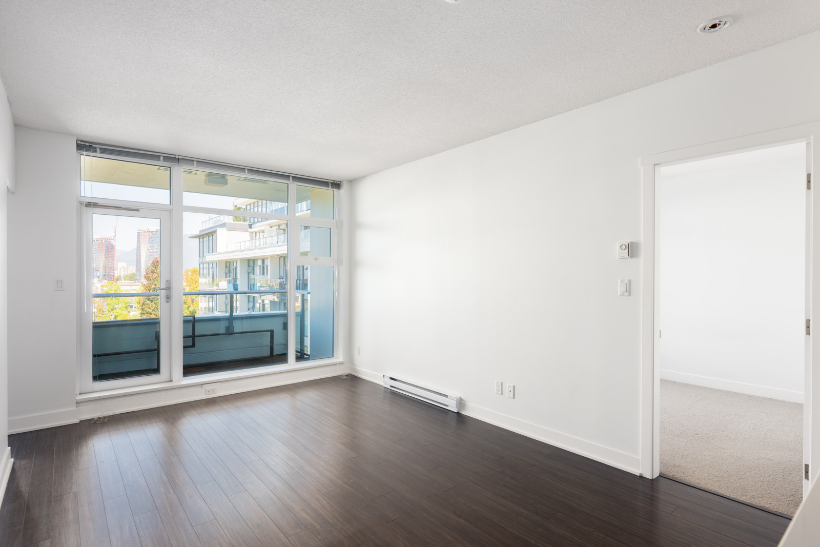 Empty modern apartment room with large windows, dark wood floors, white walls, and an open doorway leading to a carpeted bedroom.