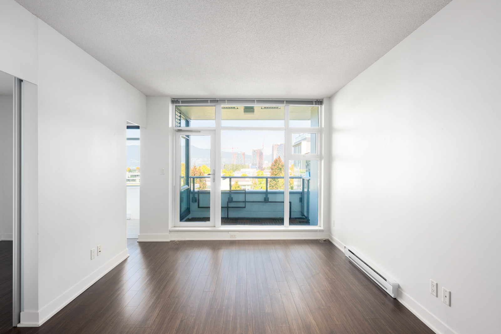 Empty room with white walls, dark hardwood floors, and large windows leading to a balcony with a city view.