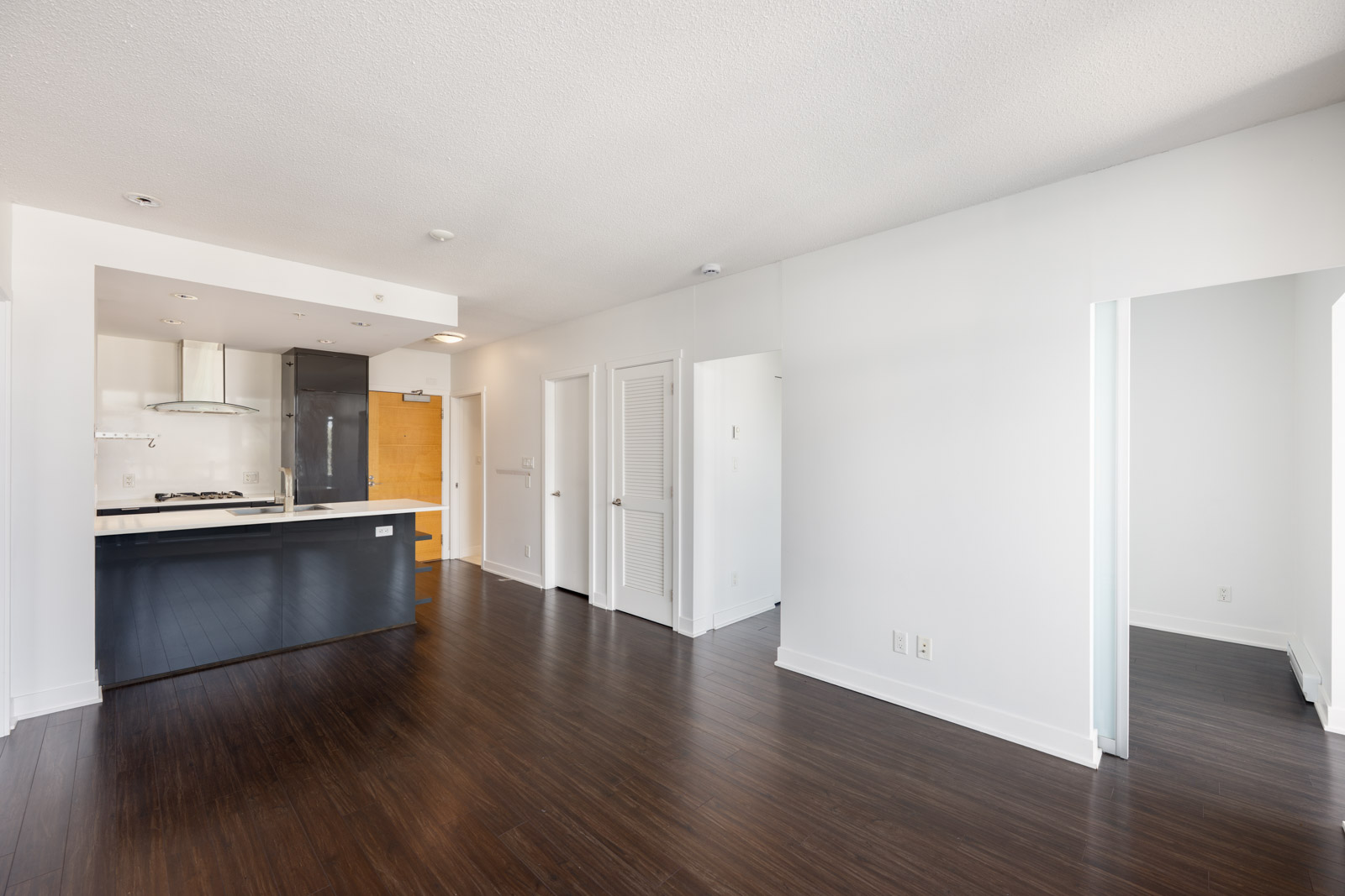 Modern, unfurnished apartment with dark wood flooring, white walls, an open kitchen with black cabinetry, and multiple doorways leading to other rooms.