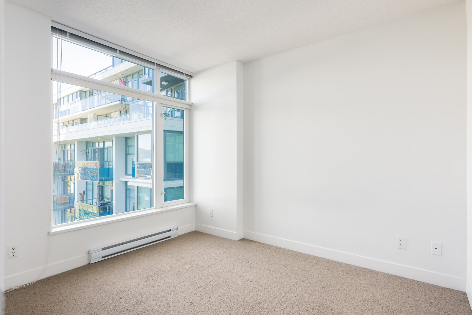 Empty room with beige carpet, white walls, and a large window overlooking a modern apartment building with balconies.