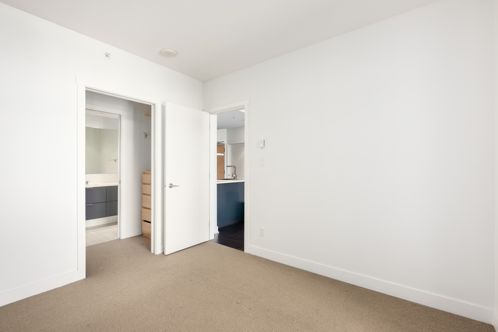 Empty room with beige carpet, white walls, and two open doors—one leading to a bathroom and the other to a hallway with a view of a kitchen area.
