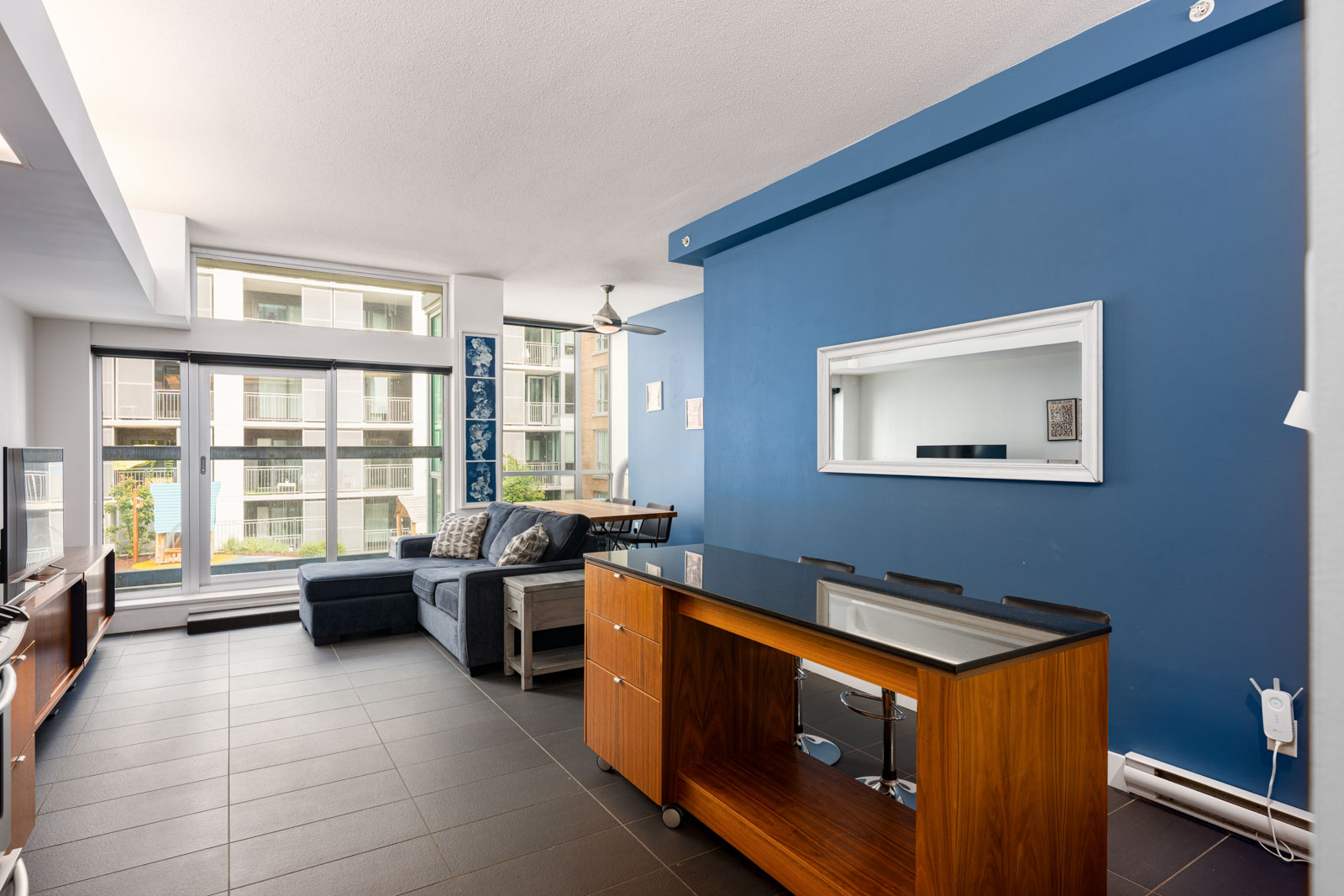Modern living room with blue accent wall, sectional sofa, wall mirror, and dining table, featuring large windows letting in natural light and view of adjacent building.