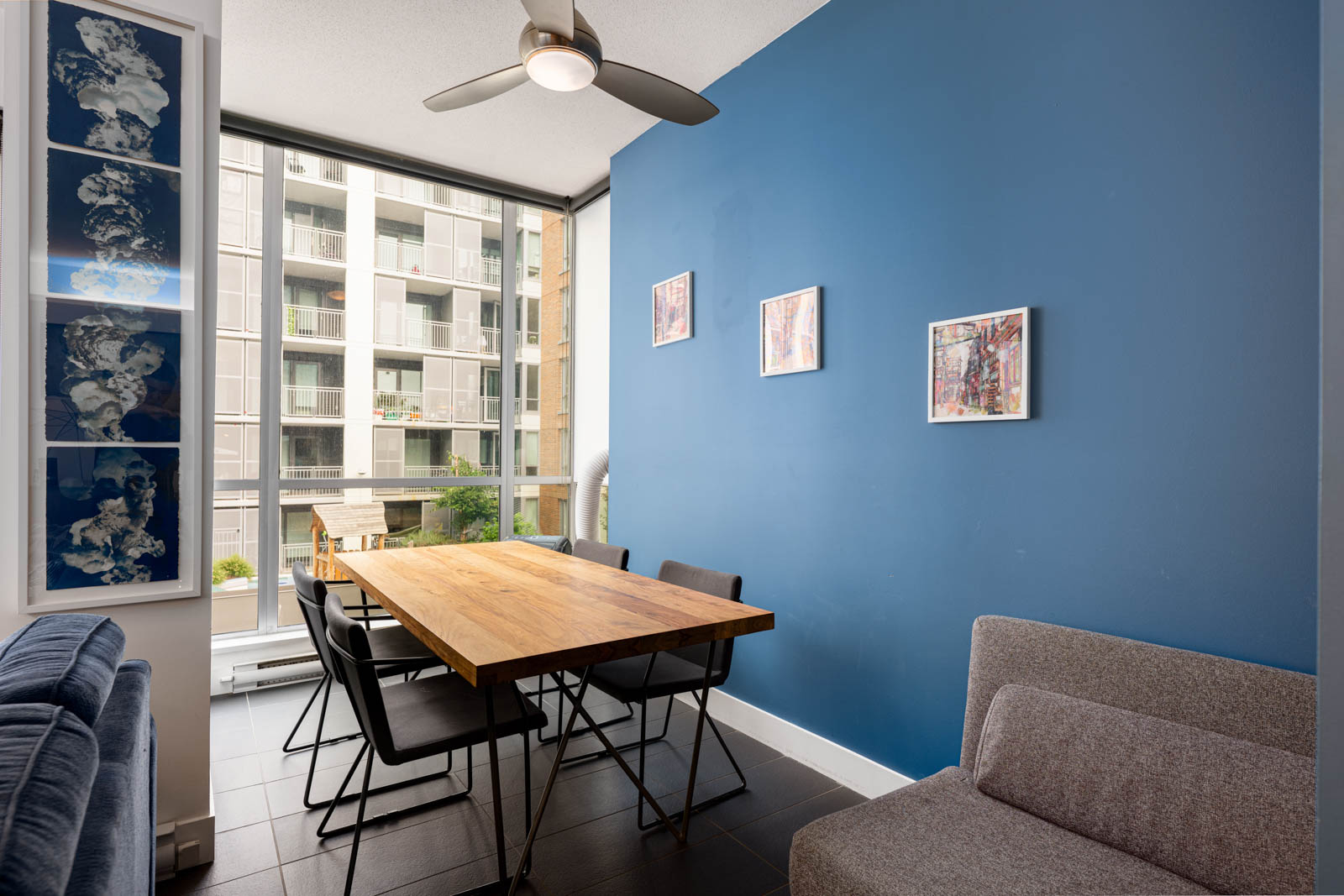 A modern dining area with a wooden table, black chairs, a blue accent wall with three framed artworks, and large windows overlooking an apartment building.