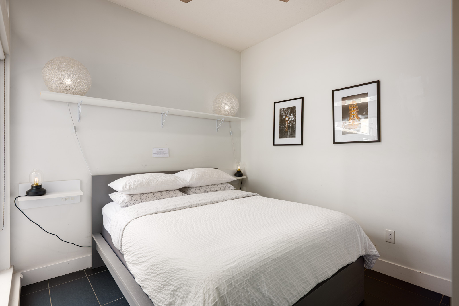 A neatly made bed with white bedding is centered in a small, bright bedroom. Two framed pictures hang on the wall, with lamps and a shelf above the bed.