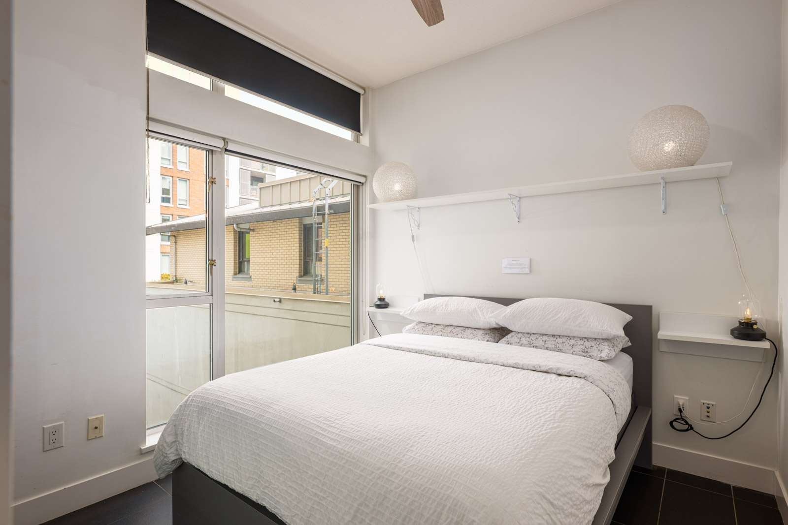 A modern bedroom with a neatly made bed, white bedding, two spherical lamps on wall shelves, large window, and neutral decor.