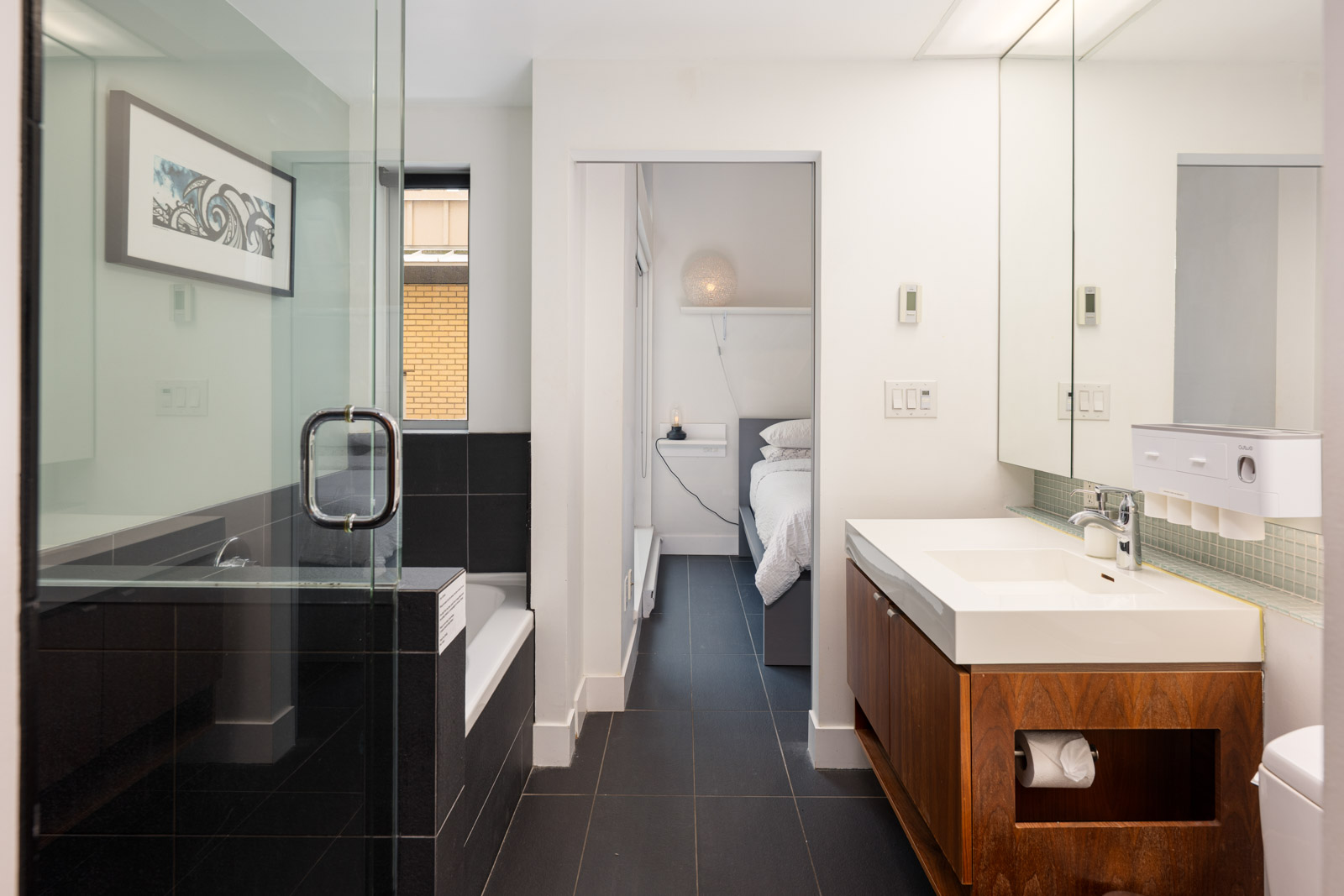 Modern bathroom with a glass-enclosed shower, white sink on a wooden vanity, and a view into a bedroom with a bed and bedside lamp.