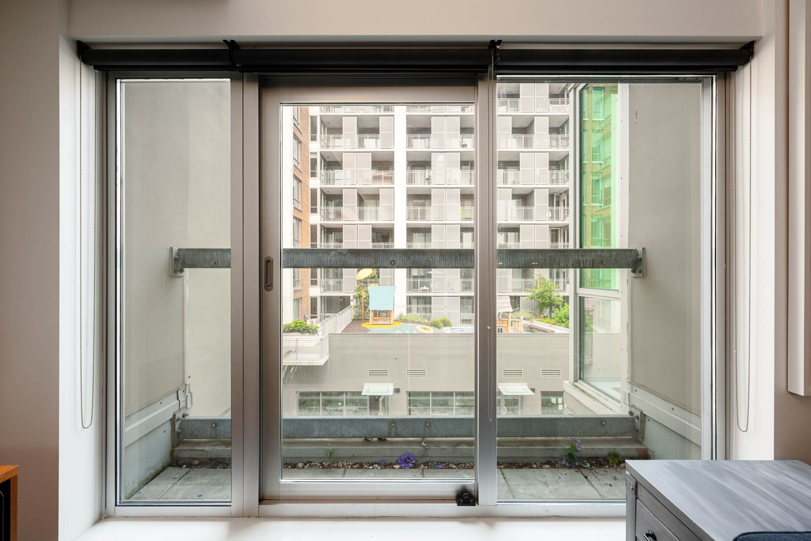 A glass sliding door opens to a narrow balcony with a metal railing, overlooking a modern apartment building across a courtyard.
