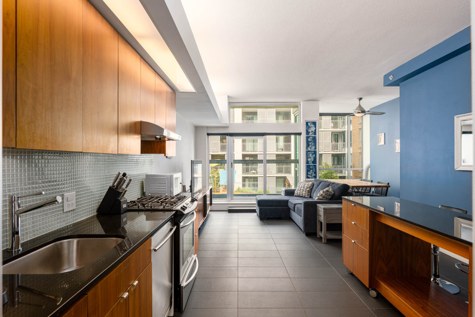 Modern apartment kitchen with wood cabinets, stainless steel appliances, and black countertops, opening to a living area with a sectional sofa and large windows.