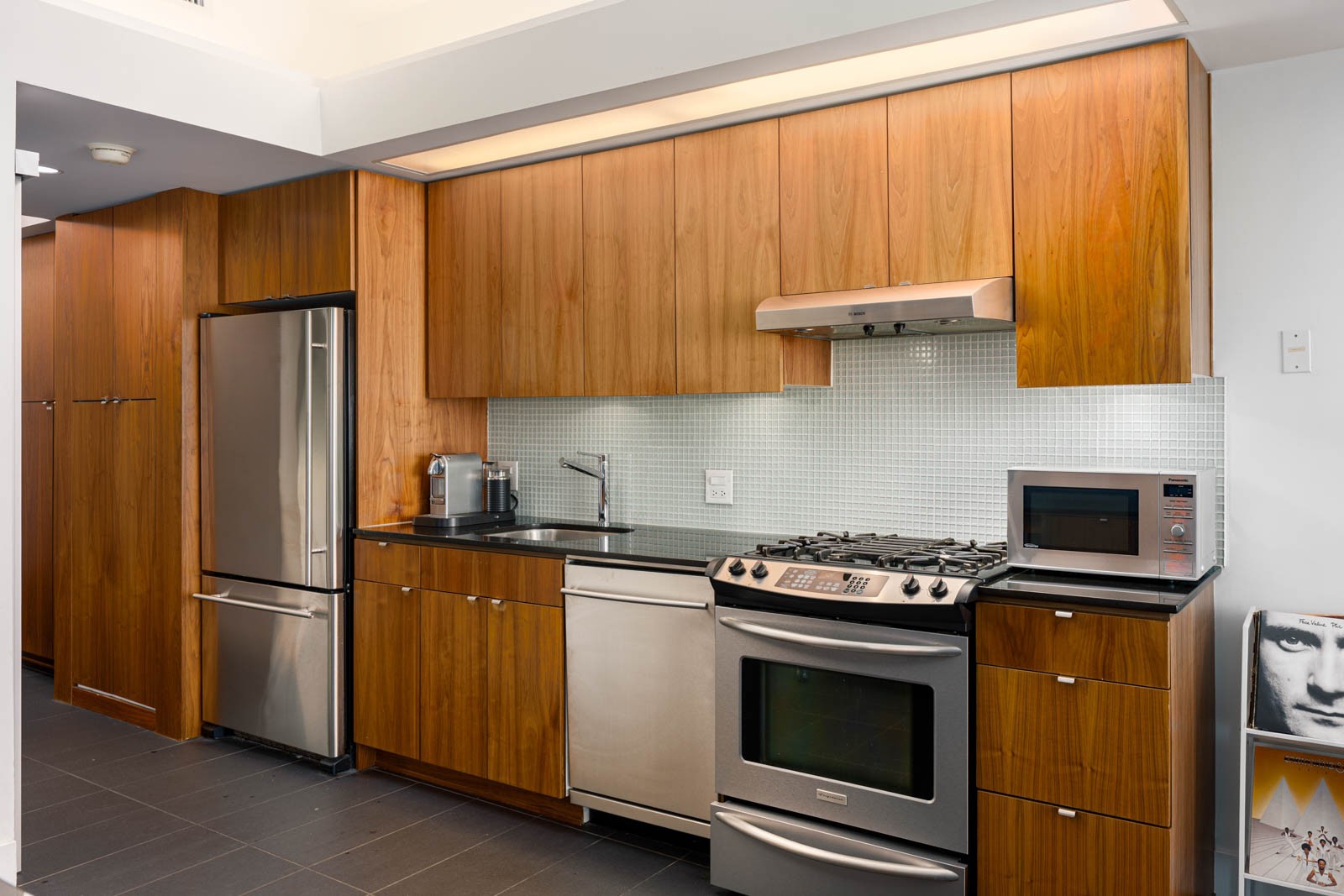 Modern kitchen with stainless steel appliances, wood cabinets, tile backsplash, and small countertop appliances; a framed portrait rests on the floor in the corner.