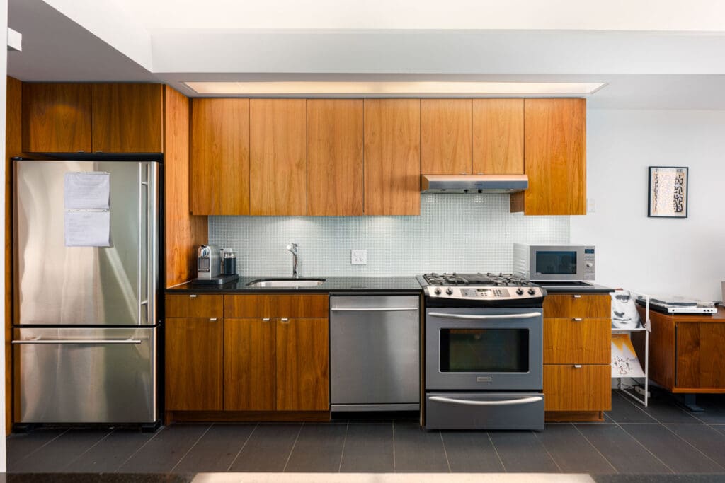 Modern kitchen with wood cabinets, stainless steel refrigerator, oven, and dishwasher, a microwave, kettle, and minimal countertop appliances.
