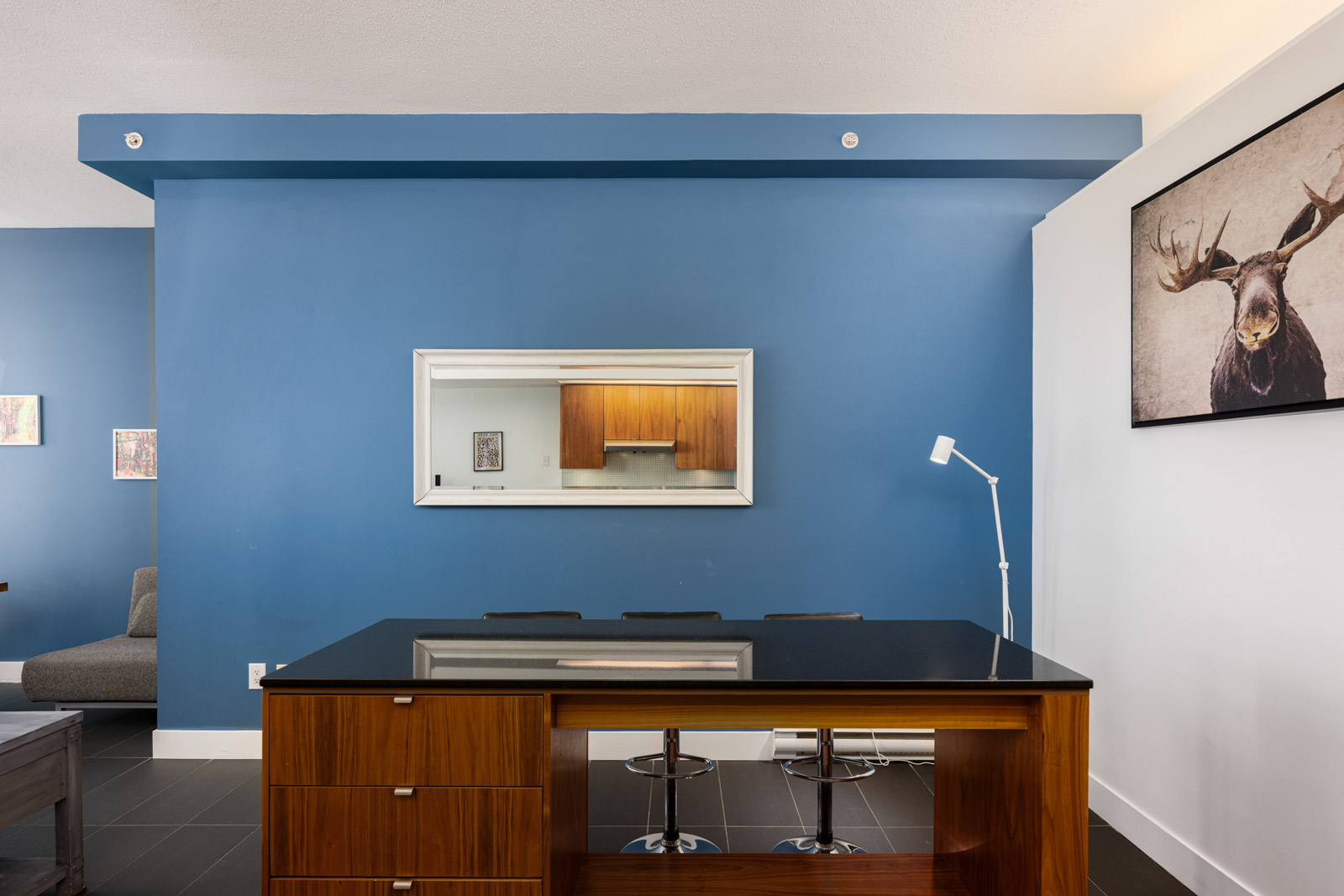 A modern home office with a wooden desk, three stools, blue accent wall, wall art featuring a moose, and a white floor lamp next to the desk.