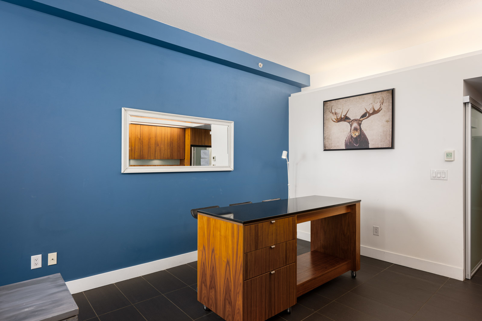 A small room with a blue accent wall, a rectangular mirror, a wooden desk, a standing lamp, and a framed moose picture on a white wall.