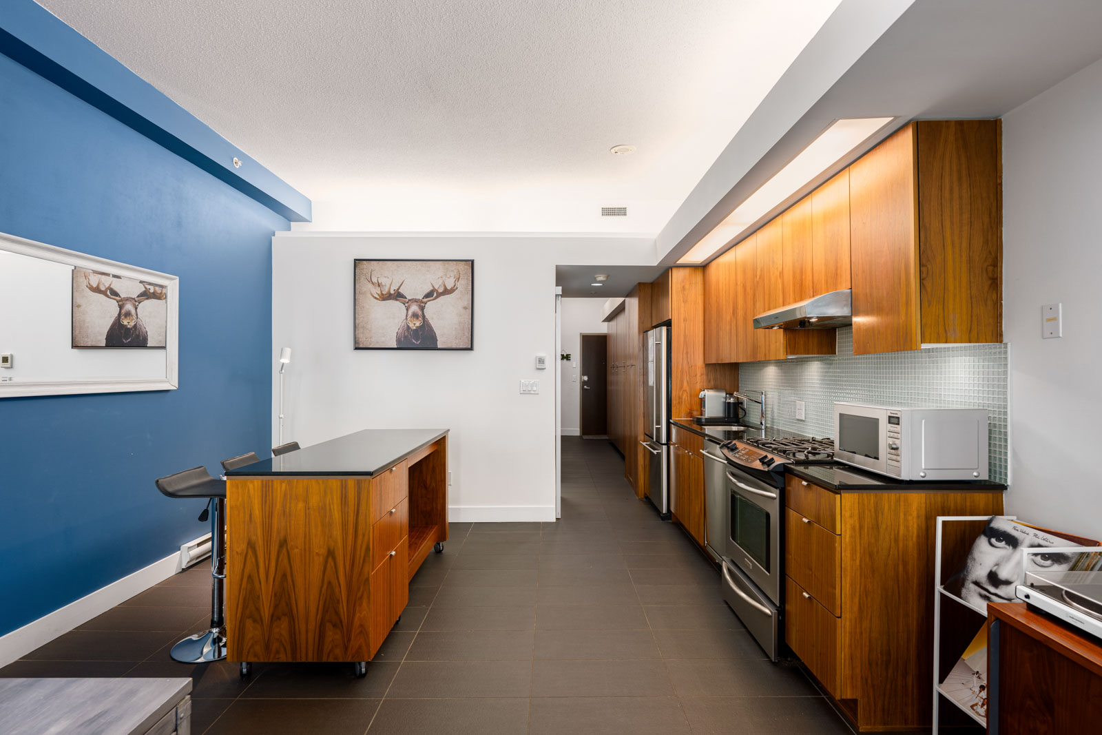 Modern kitchen with wood cabinets, stainless steel appliances, a blue accent wall, island with two stools, and moose artwork above a small table.