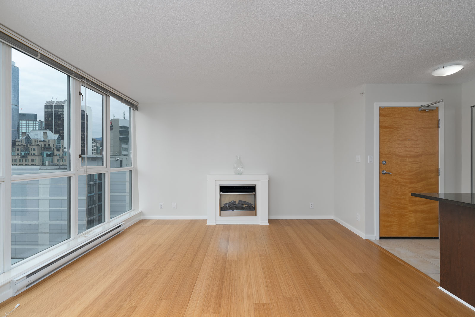 A modern, unfurnished apartment living room with large windows, city views, wood flooring, a small fireplace, and a wooden entry door.