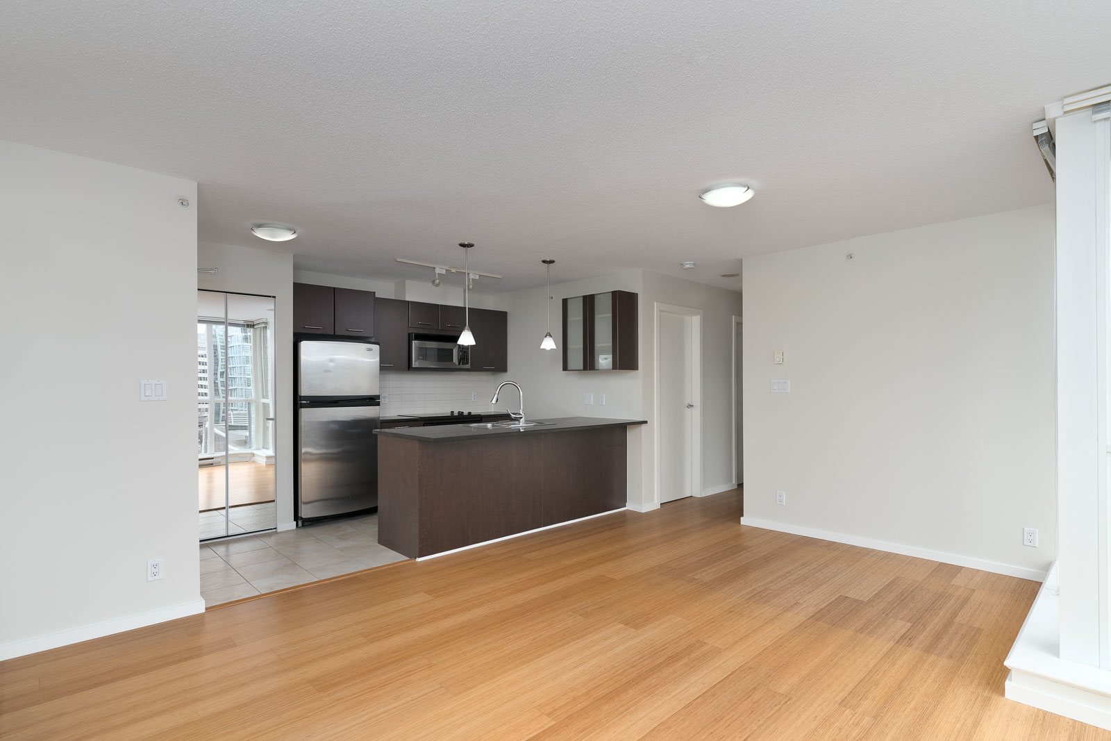 Modern apartment interior with an open kitchen featuring stainless steel appliances, wood cabinets, an island, and a spacious living area with wood flooring.