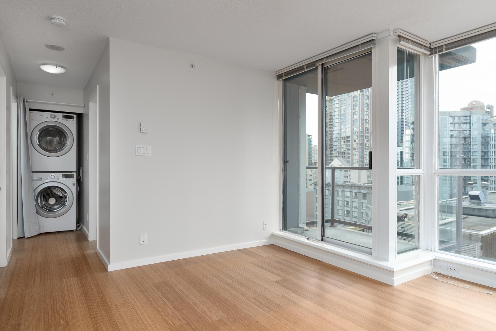 A modern apartment room with light wood flooring, large windows with city views, and a stacked washer and dryer in a hallway alcove.