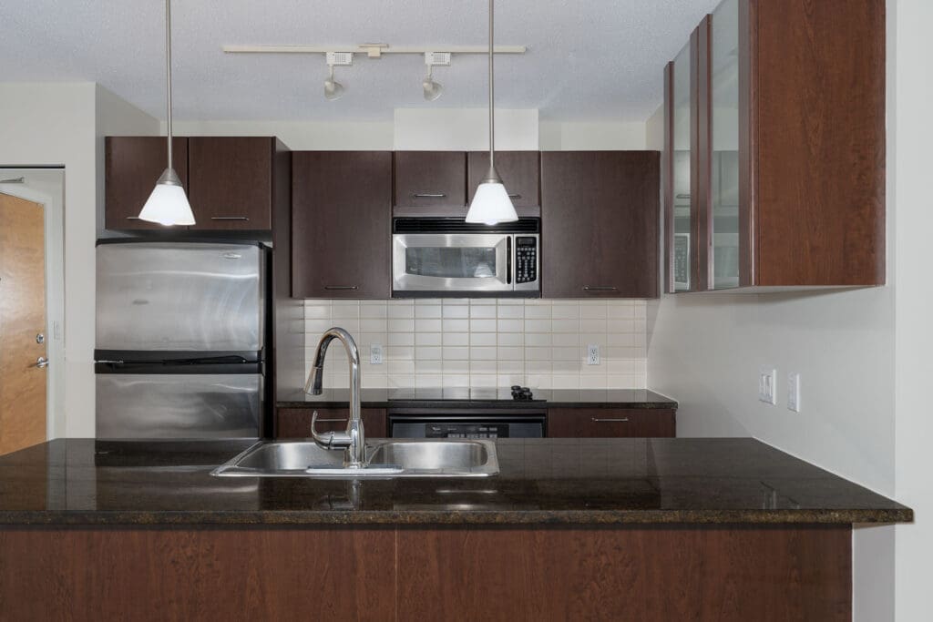 Modern kitchen with dark wood cabinets, stainless steel appliances, double sink, black countertop, and pendant lighting above an island.