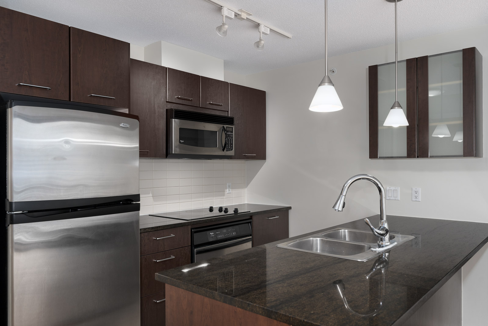 Modern kitchen with dark wood cabinets, stainless steel appliances, black countertop island with sink, white tile backsplash, and pendant lighting.