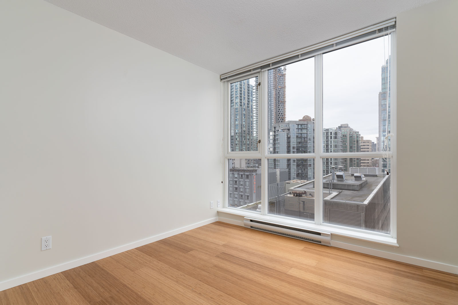 Empty room with light-colored walls, hardwood floors, and large windows overlooking a cityscape with tall buildings.