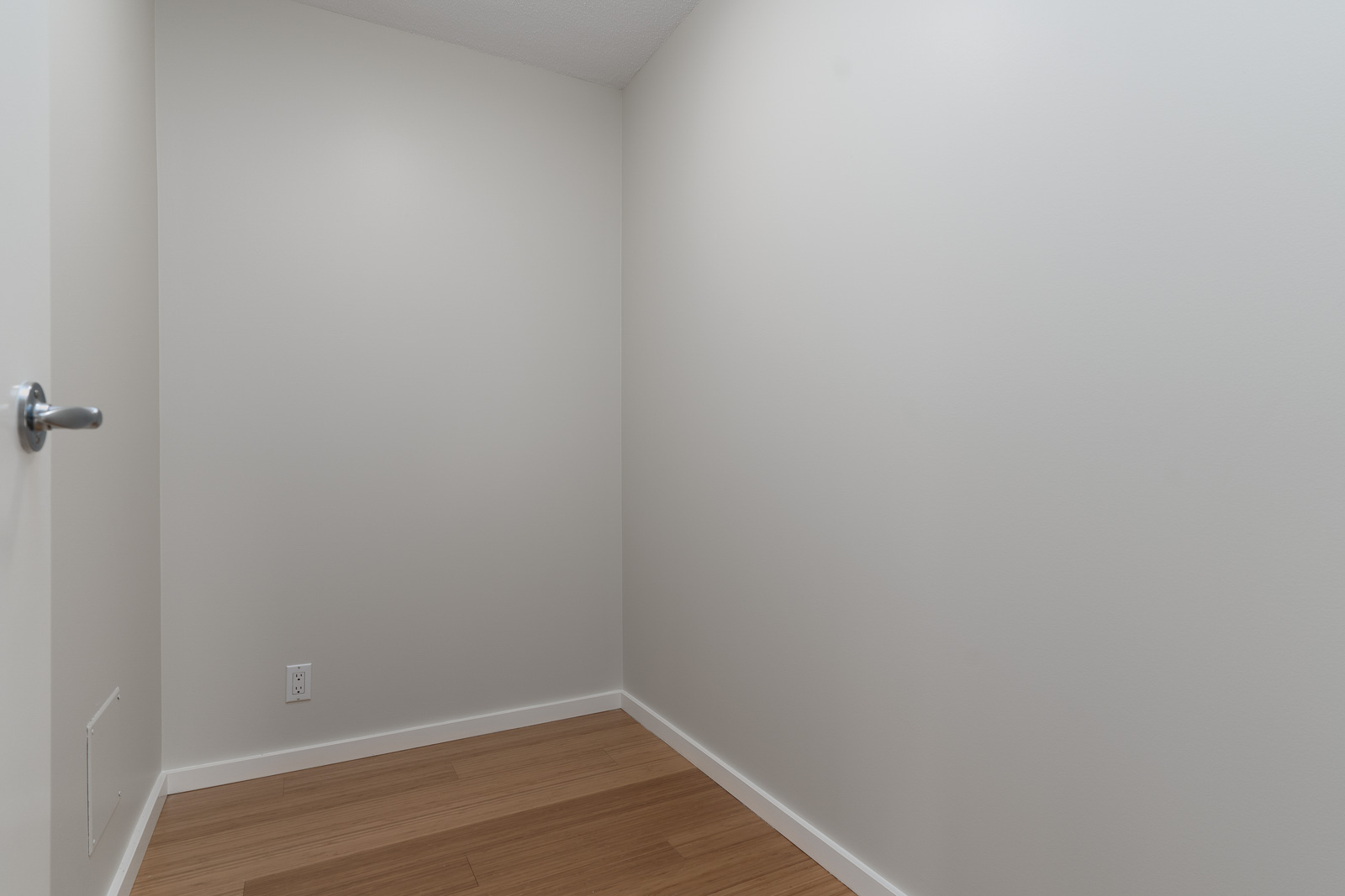 Small, empty room with plain white walls, a closed door on the left, wooden floor, and a single electrical outlet near the corner.
